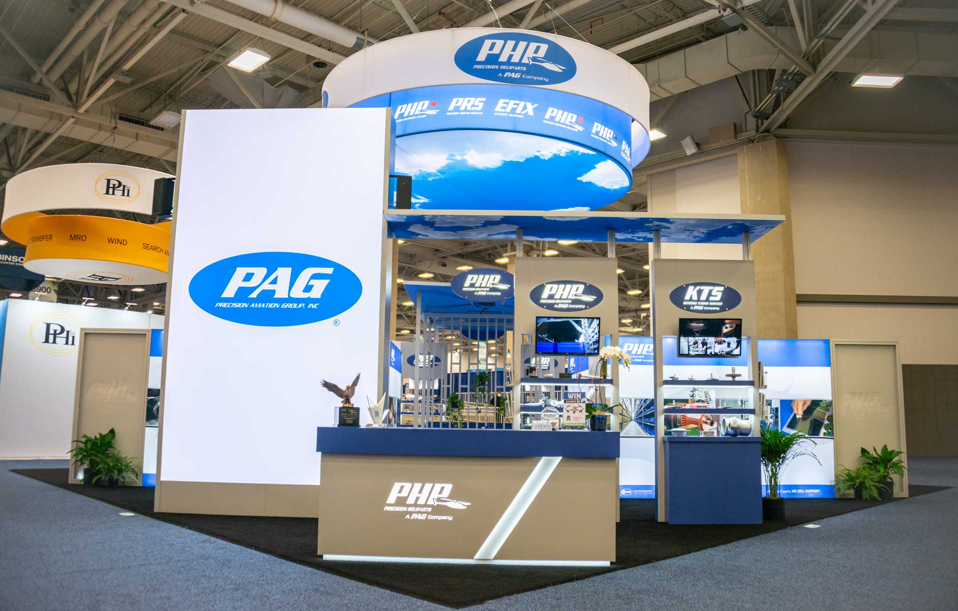 A trade show booth for "PAG," featuring a sleek, modern design with blue and white colors. The booth has a reception counter, displays with product images, and digital screens. The PAG logo is prominent, with a circular overhead sign and banners.