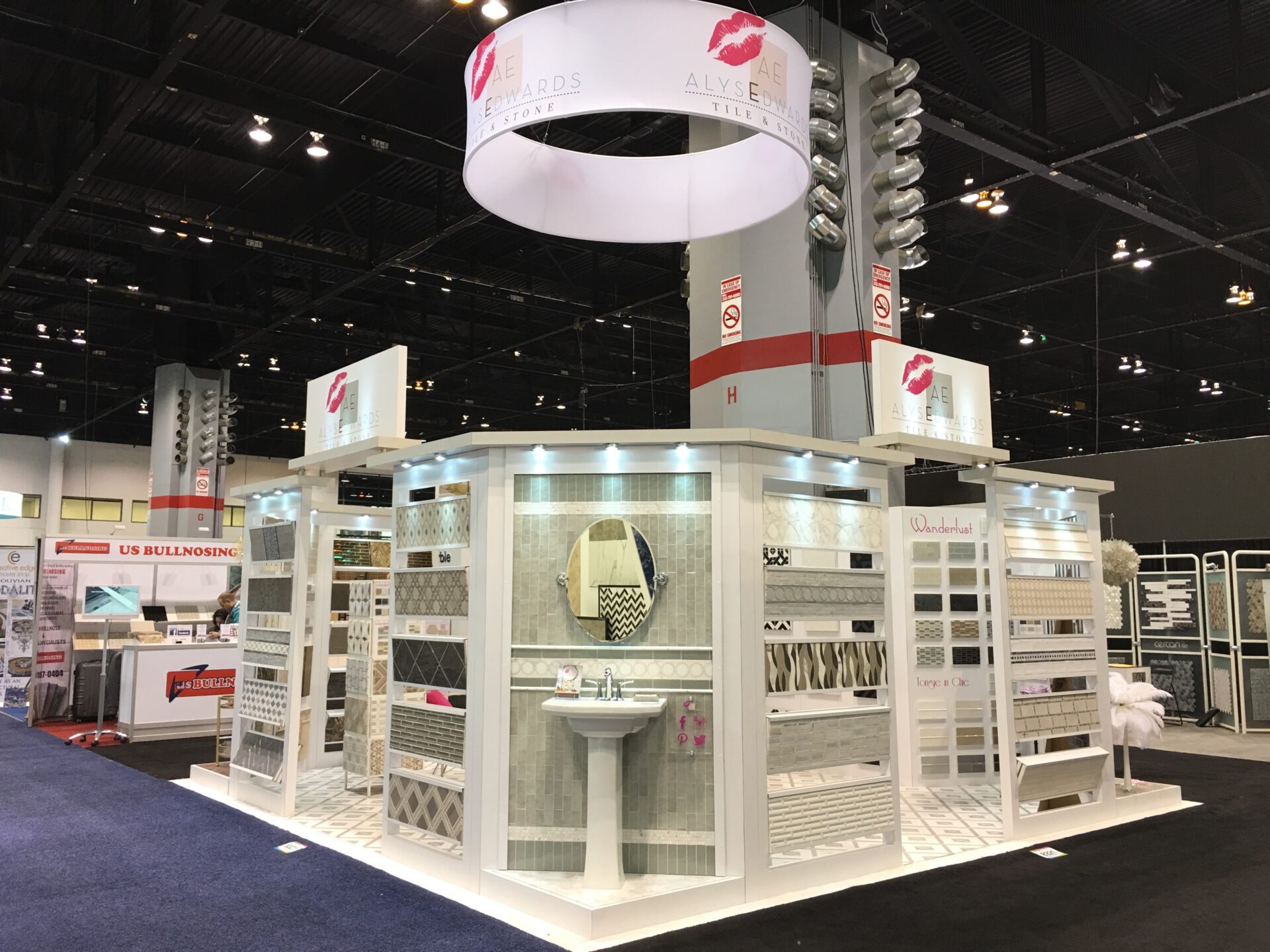 Trade show booth featuring a white display structure showcasing various tile samples. The booth has a semi-circular top banner with logos, a central washbasin with a decorative mirror, and surrounding panels with different tile designs. Overhead lights illuminate the display.