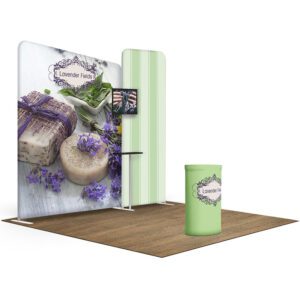 A trade show booth featuring a green cylindrical stand and a backdrop with images of lavender-themed soap and sprigs of lavender. The backdrop displays the text "Lavender Fields." A small display screen is mounted on the backdrop, alongside a small shelf.