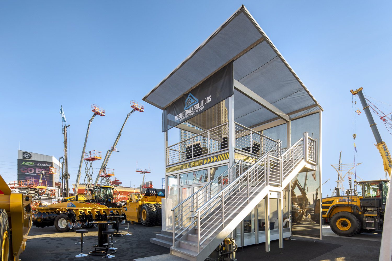 An industrial building with two levels, featuring a staircase on the right and a sign that reads "McLaren Solutions". Surrounding the building are large construction vehicles and equipment, with cranes and machinery in the background against a clear blue sky.