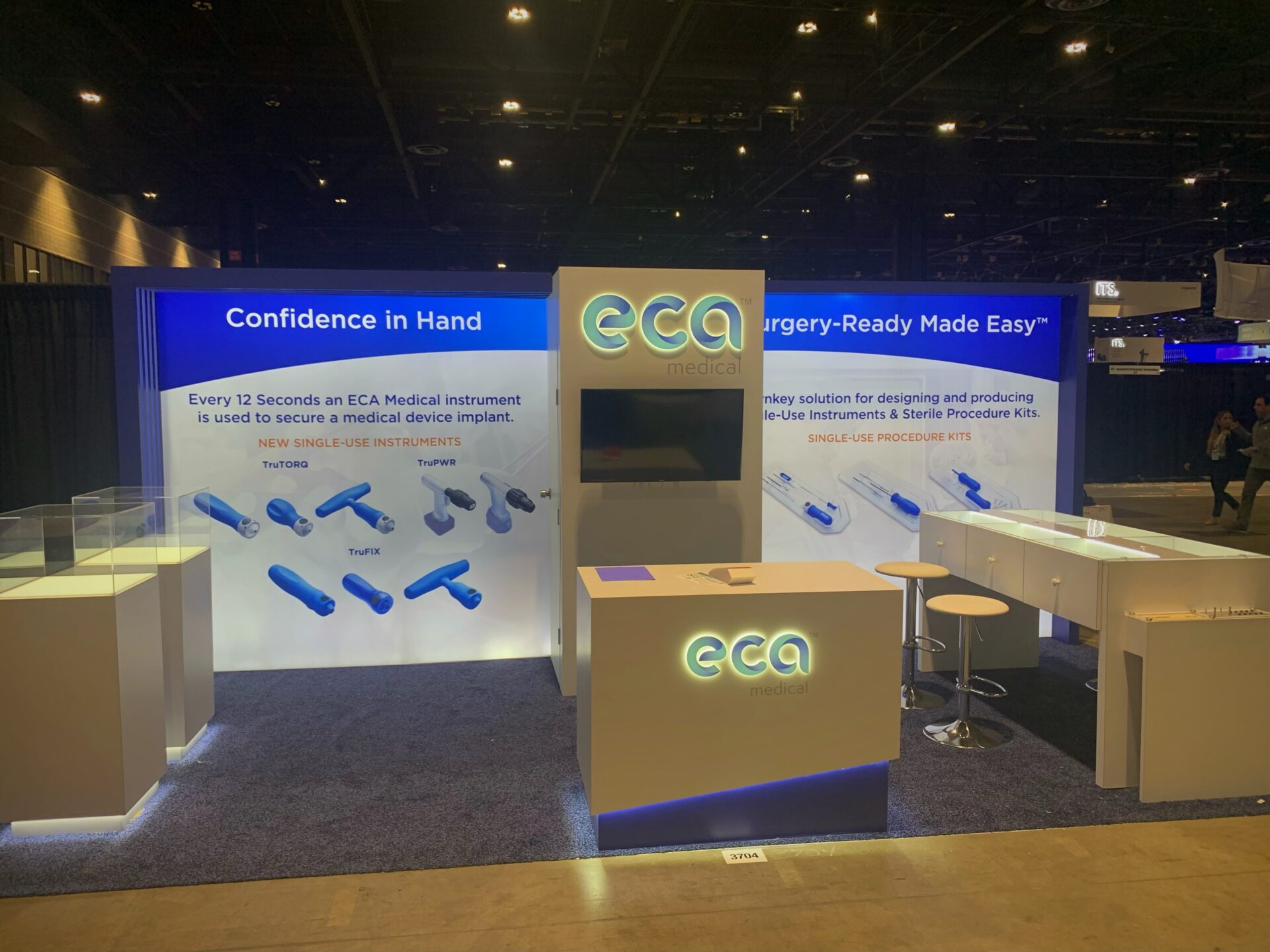 A trade show booth for ECA Medical is displayed, promoting medical instruments with two slogans: “Confidence in Hand” and “Surgery-Ready Made Easy.” The booth has a central TV screen, counters with flyers, and lit-up signage featuring the ECA Medical logo.