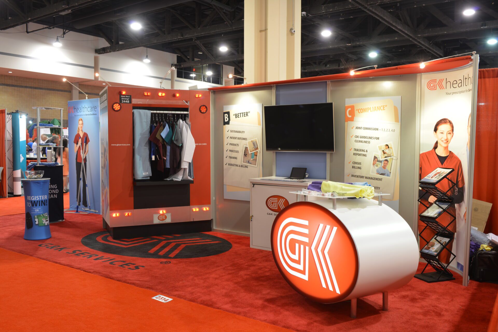 A trade show booth displays a company named G&K Services. The booth features a rotating clothing rack, a mounted TV, branded signage, and a reception counter with a company logo. Brochures are in a rack to the right, and another booth is visible in the background.