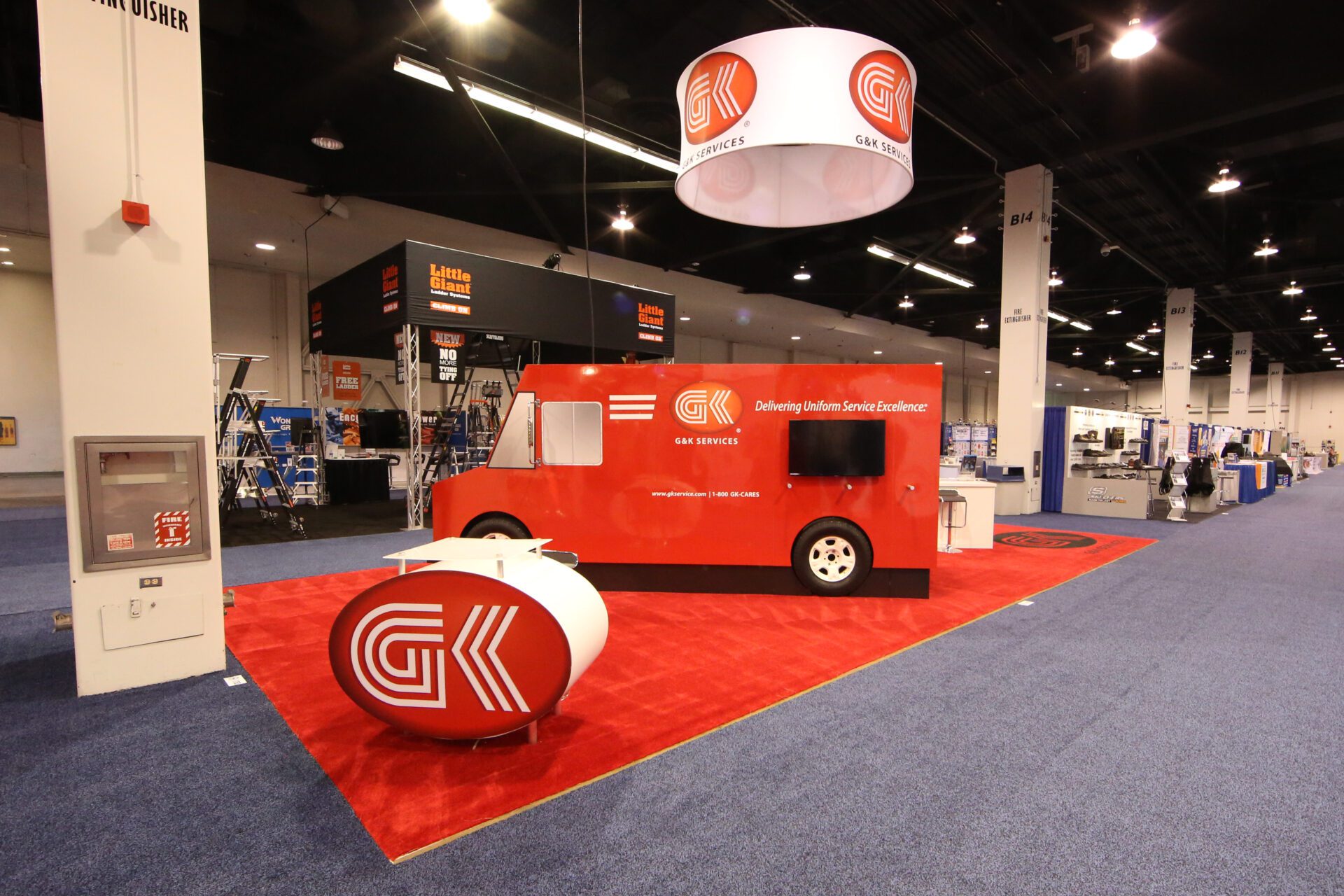 An exhibition booth with a red truck, branded "GK" and "GAI Services," features a red carpet, circular table, and overhead signage. The booth's theme is "Delivering Uniform Service Excellence," set in a well-lit convention center, with other displays in the background.