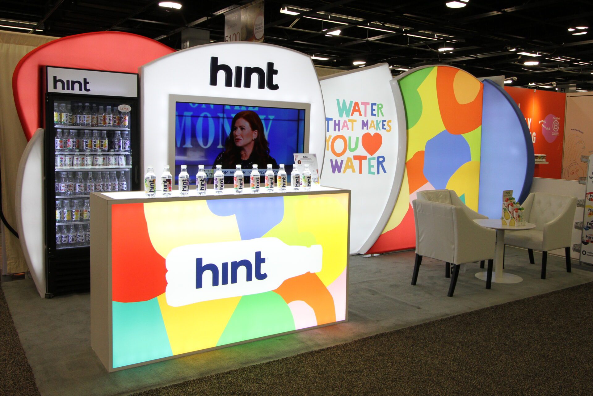 A brightly lit booth showcases Hint products at a convention, featuring a colorful display with a prominent "Hint" logo. Bottled Hint water is arranged on a counter. Behind the counter, a screen shows a woman speaking. A refrigerator and a "Water that makes you water" sign are also visible.