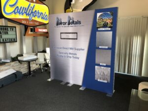 A trade show booth with a banner that says "Mirror Metals, The Source for Specialty Sheet Metal Finishes." It features samples of four different metal finishes labeled "Bright Mirrored," "Embossed," "Etched," and "Pattern." The area also has vintage signs.