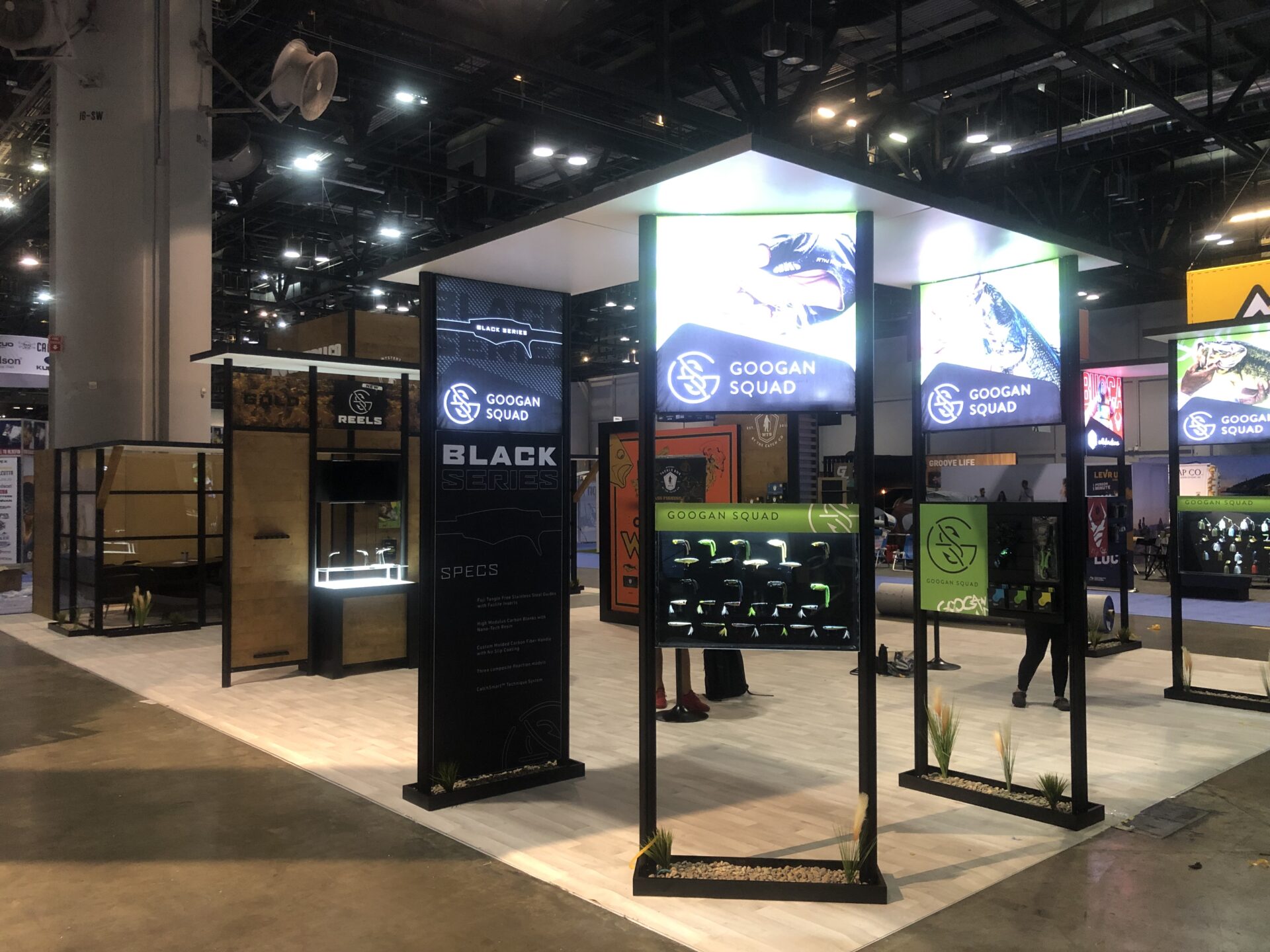 A well-lit trade show booth features the Googan Squad, showcasing their fishing gear. The booth has illuminated signs, display racks with products, and informational panels. The setup includes interactive displays and promotional graphics under a high ceiling.