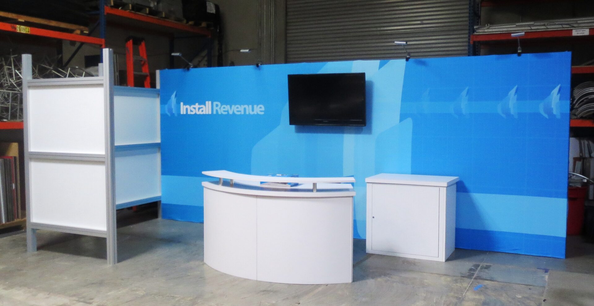 A blue trade show booth with a large backdrop displaying the text "Install Revenue." The setup includes a curved table, a tall display stand with empty white panels, a flat-screen TV mounted on the backdrop, and a small white cabinet.