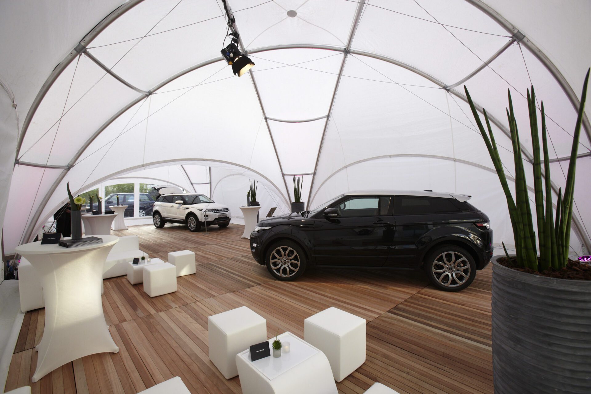 A modern, white exhibition tent features a wooden floor, minimalist white furniture, and plants. Inside are two luxury SUVs, one black and one white. Pedestals with displays and lighting fixtures are also visible, creating a sleek, contemporary atmosphere.