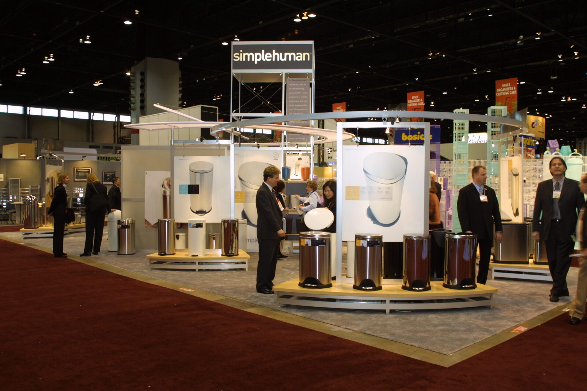 A trade show exhibit features 'simplehuman' branded products, including trash cans displayed on elevated platforms. Several attendees examine the items, with the exhibition hall filled with booths in the background. The carpet is maroon-colored.