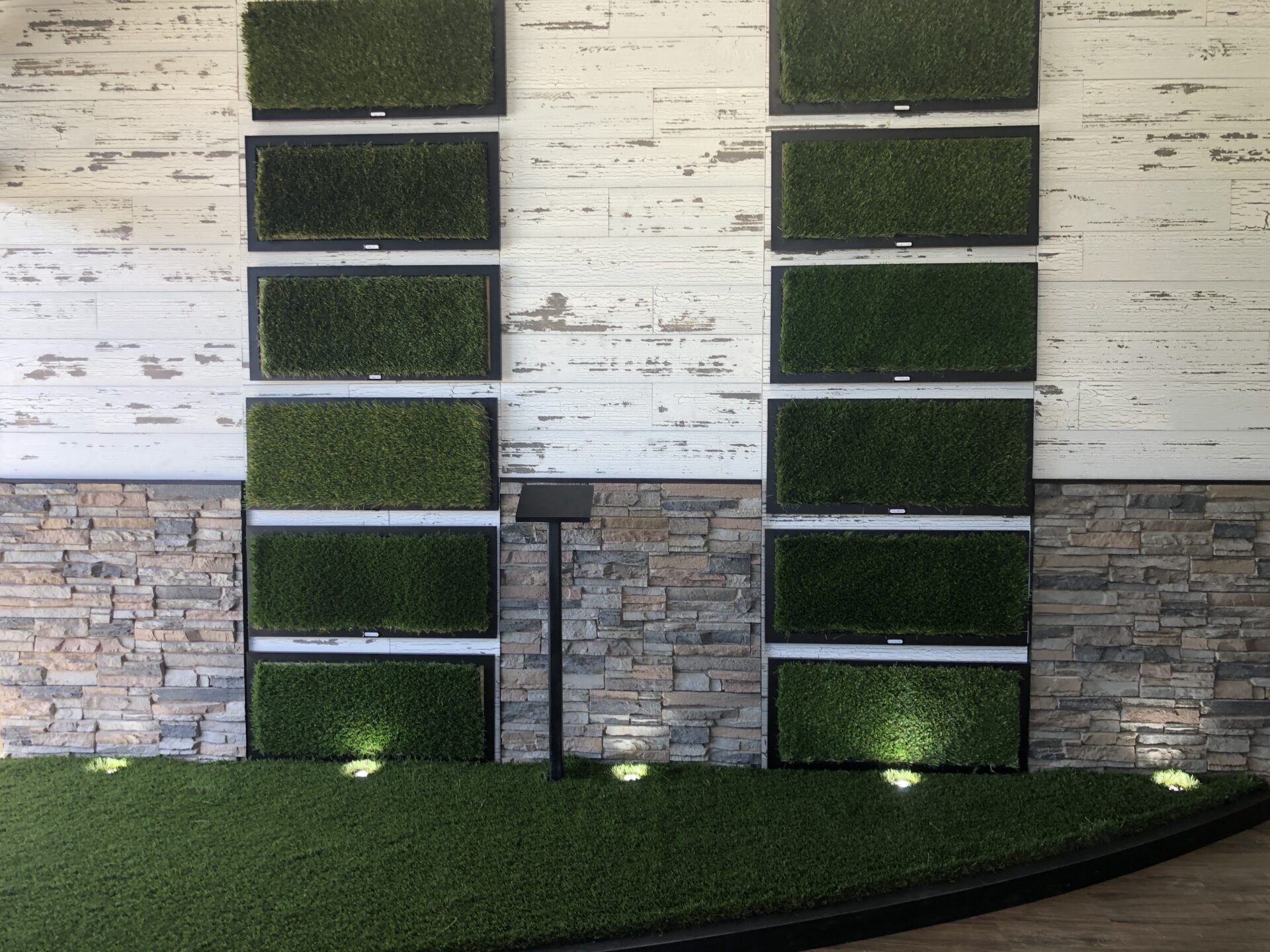 A display featuring vertical panels of green artificial turf against a white wall with horizontal wooden planks. The lower half of the wall is covered with stone veneer. Circular lights illuminate the green turf below the panels.