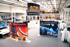 A large industrial room with high ceilings, showcasing three booths with banners labeled "ATM Fly-Ware," "Rigging Solutions," "Speaker Mounting & Rigging," and "Mounting Solutions." The space contains various equipment, tables, and tools, with a mezzanine in the background.