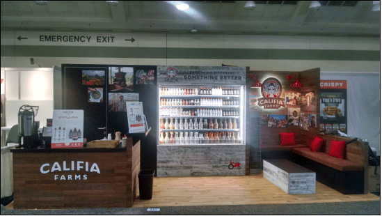 A trade show booth featuring Califia Farms products. A counter with samples and promotional materials is to the left, while a well-lit display of various bottled beverages is centered. Red cushioned seating and informational posters are arranged around the booth.