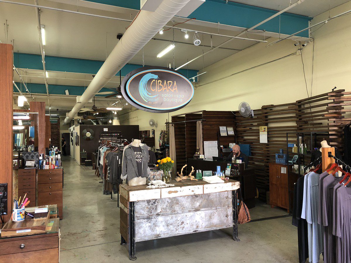 A modern, well-lit salon and spa boutique with wooden decor. There are clothing racks, a display table with folded garments, and shelves with beauty products. The sign above reads "CIBARA Salon Spa Boutique." A staff member is seen working at the reception desk.