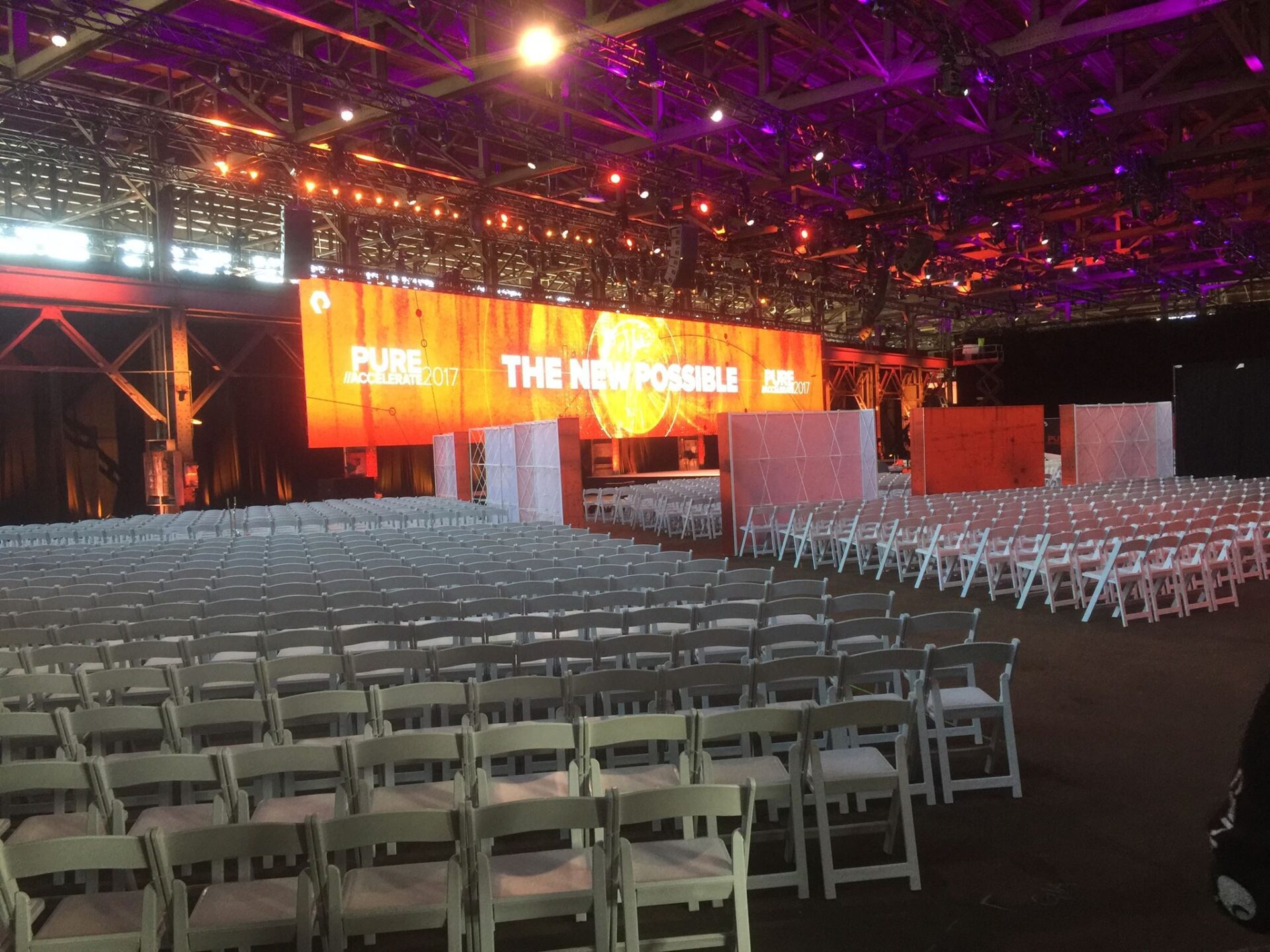 A large indoor event space is set up with rows of white chairs facing a stage with a large, illuminated screen. The screen shows text that reads "PURE//ACCELERATE 2017 - THE NEW POSSIBLE." Purple and orange lighting creates a vibrant atmosphere.