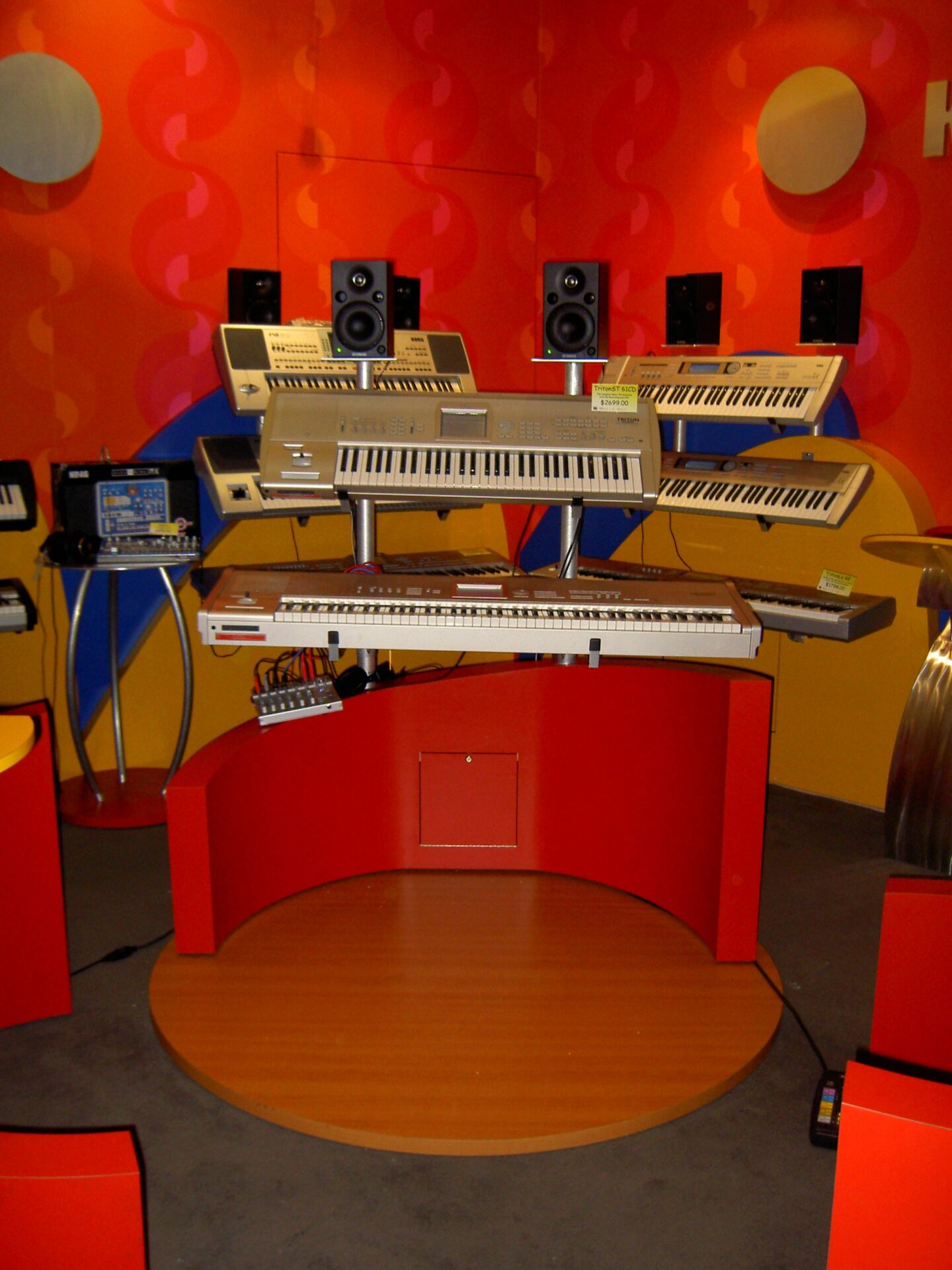 A vibrant music studio setup featuring multiple keyboards on stands, surrounded by speakers and electronic equipment. The room has bright orange and red walls with curved patterns, creating an energetic atmosphere.