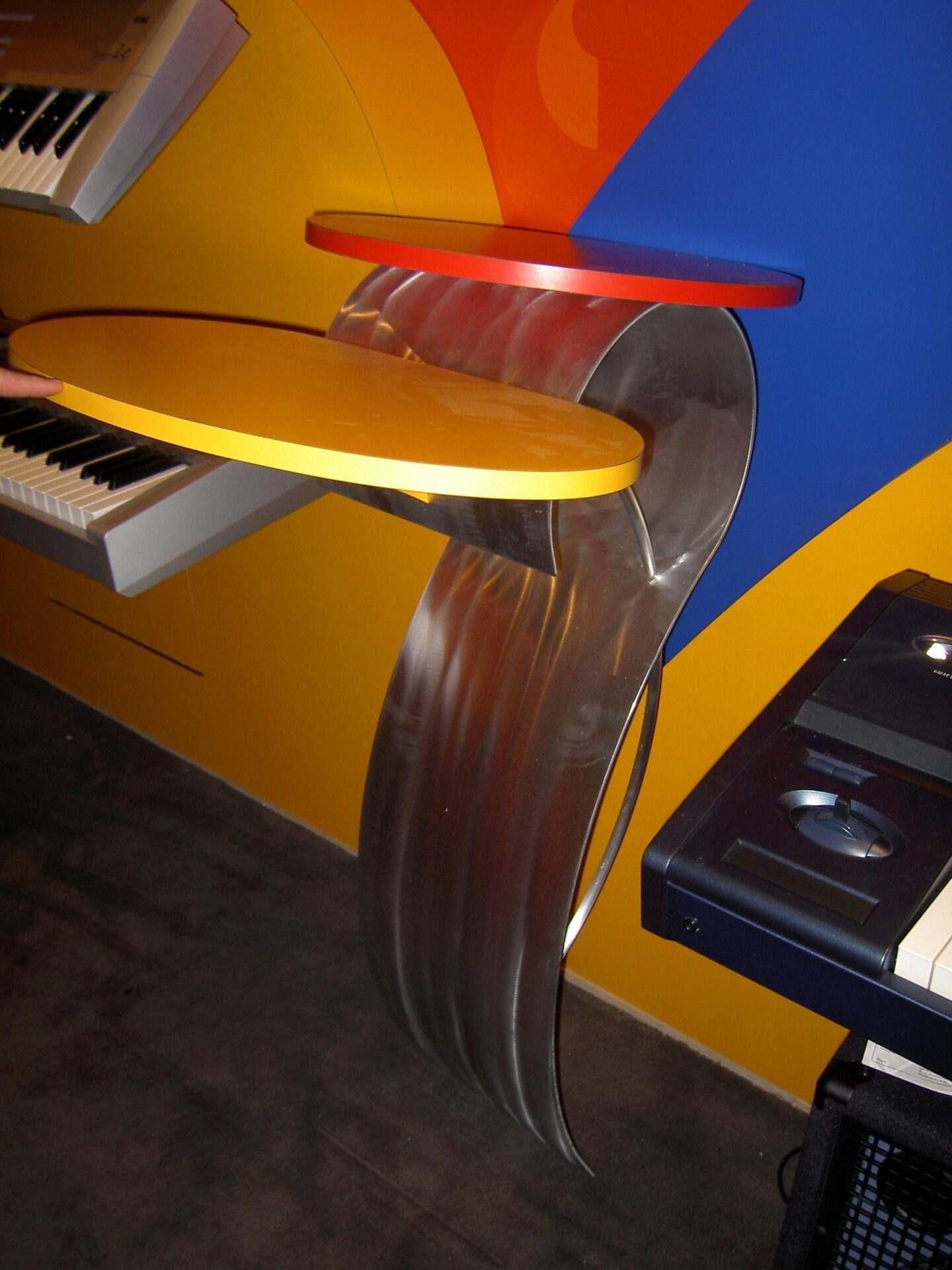 A modern music workstation setup with sleek, colorful design elements. It features a curved metallic structure supporting two layered platforms in yellow and red. Nearby are electronic keyboards mounted on an orange and blue wall. The area has a dark carpeted floor.