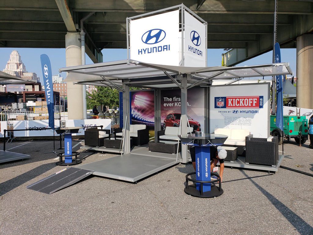 Outdoor Hyundai display area at an event. The setup includes seating with white cushions, a Hyundai logo banner, and NFL Kickoff promotional signs. It sits under an overpass, with a cityscape and various sponsor flags in the background.