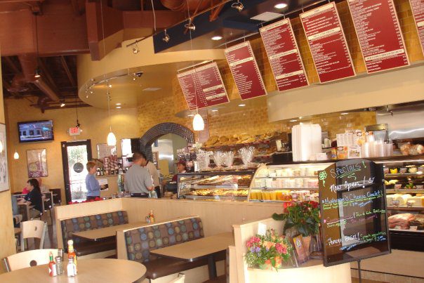 A cozy café with light wooden furniture and a colorful menu board above the counter. There are customers ordering at the counter, which displays various pastries and baked goods. Potted plants, tables, chairs, and a small blackboard with specials create a welcoming atmosphere.