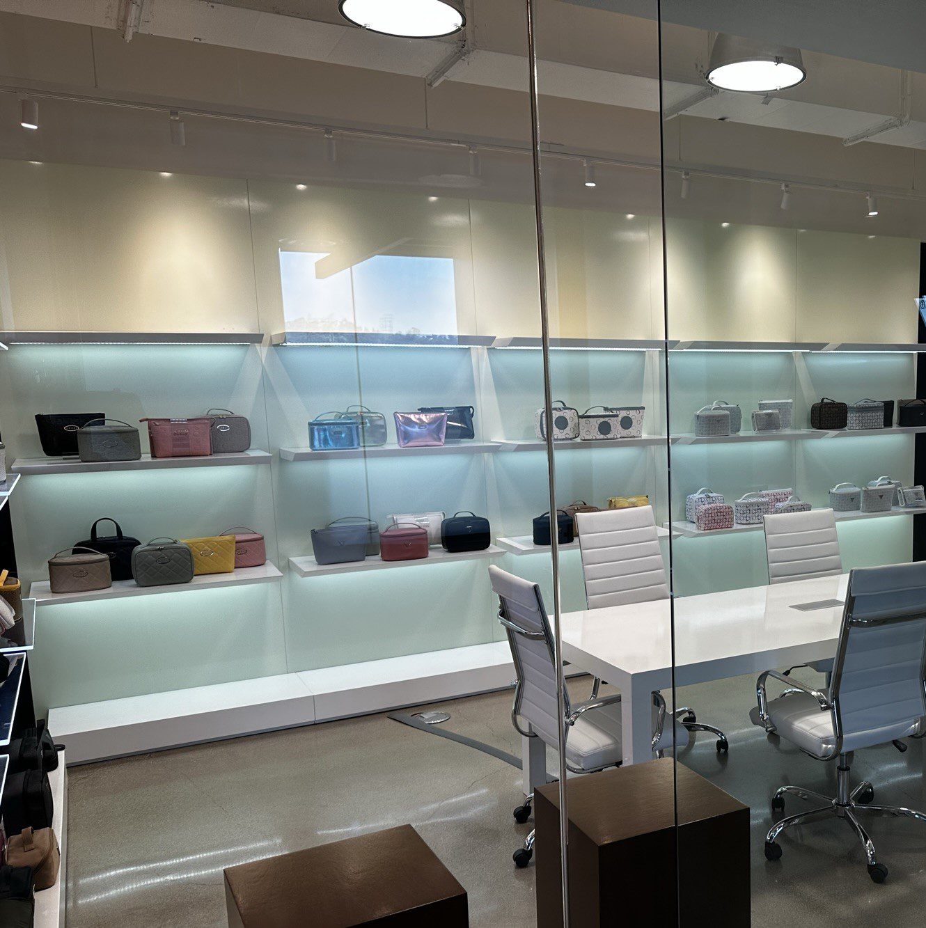 A modern retail store displays handbags on illuminated shelves. The space is brightly lit with white chairs around a table, giving an impression of a chic showroom. The handbags vary in colors and designs, neatly organized on the back wall.